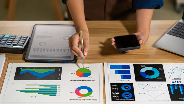 Business professional reviewing charts at a desk, pointing to colorful graphs while holding a phone in a modern office, showing planning, analysis, and financial decision making.