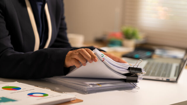 Businesswoman reviewing a stack of documents at an office desk with charts, binder clips and laptop, showing paperwork, analysis and everyday administrative work.