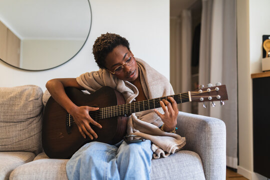 Young Woman Playing Acoustic Guitar Wrapped in Blanket on Sofa