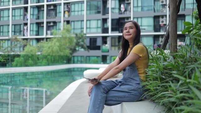  A happy young woman in casual wear sitting by the swimming pool of a modern condominium, enjoying her urban lifestyle and relaxing during a bright day in the city. 