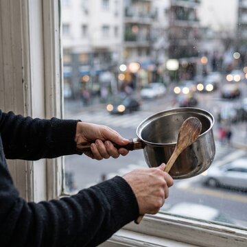 A person holds a stainless steel pot and a wooden spoon by an open window, performing a cacerolazo protest against a blurred city street background with traffic and evening lights