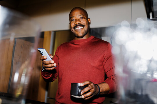 Man smiling holding smartphone and coffee mug at apartment