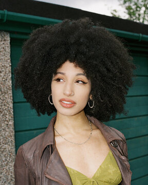 woman with afro leant against a garage looking away from the camera.