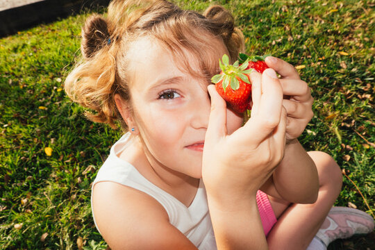 Girl holding strawberry close to face looking at camera outdoors
