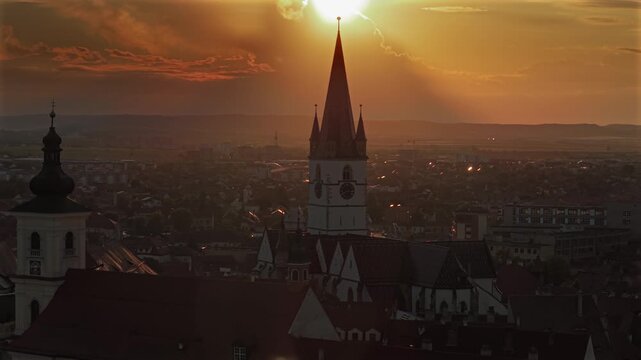 Aerial 4k video of the old town of Sibiu featuring historic red roofs and the tall church tower, with the majestic blue Fagaras mountains visible on the horizon. Sibiu, Transylvania, sunset landscape.