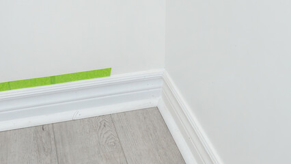 Fototapeta premium Corner of a room with freshly painted white walls, wooden floor, and green painter's tape applied along the baseboard for clean edges