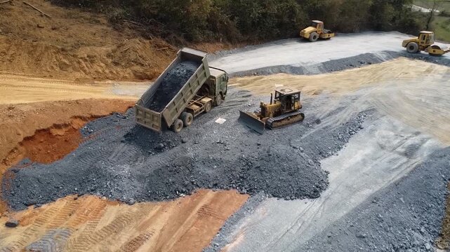 Footage of highway construction equipment operating in mountainous terrain, featuring a dump truck unloading crushed stone base material for road foundation preparation. 