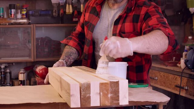 Carpenter service. Furniture making. Male joiner in gloves covering plank surface with brush glue in workplace with professional tools.