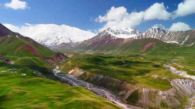 Aerial view of the lush green Alay Valley with a winding river and snow-capped Pamir Mountains under a blue sky, Alay District, Osh Region, Kyrgyzstan.