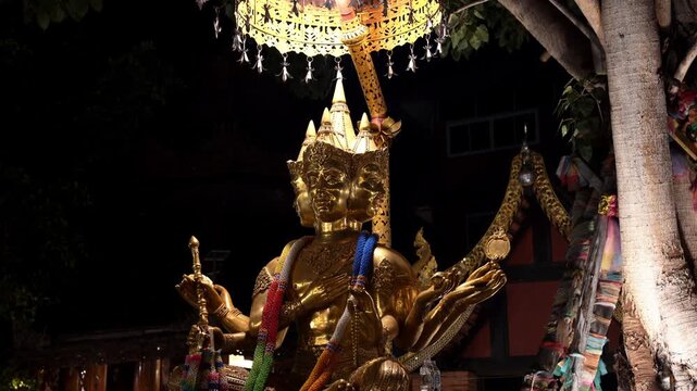 Static medium shot of Phra Phrom, the four-faced golden Brahma deity, adorned with colorful garlands and ceremonial umbrella, dramatically lit against dark night sky at a Thai shrine