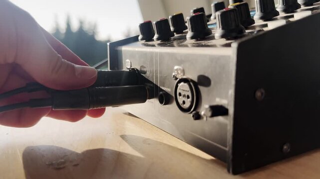 Close-up of a hand plugging a cable into a broadcast intercom audio unit, with XLR socket, control knobs, sunlight, and live production equipment detail.