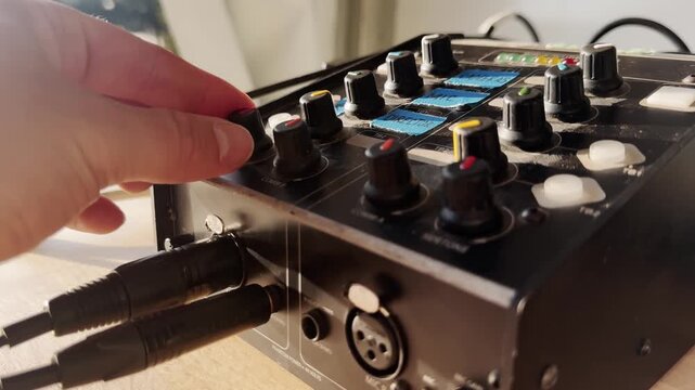 Close-up of a hand adjusting a comms knob on a broadcast intercom unit, with connected audio cables, control dials, sunlight, and live production gear.