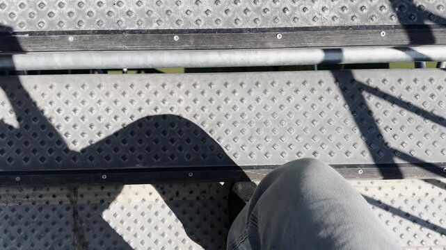 Point-of-view shot of a person walking on a perforated metal platform in stadium stands, with shadows, black shoe, railing lines, and sports venue walkway detail.
