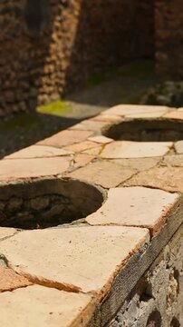 Vertical close-up of a Pompeii thermopolium counter with circular dolia openings, cracked tile surface, warm sunlight, and blurred ancient stone walls.