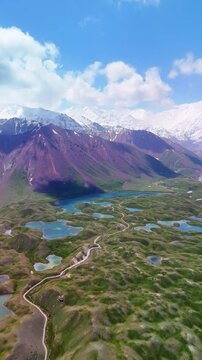 Aerial view of Tulpar Lake and a winding road through green hills and purple mountains under snow-capped peaks in Alay District, Osh Region, Kyrgyzstan.