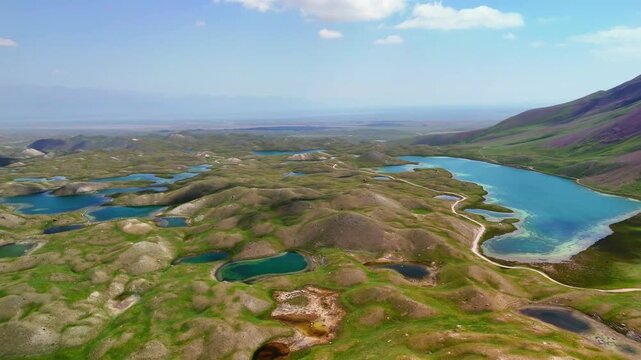 Aerial view of Tulpar Lake surrounded by rolling green hills and the majestic Pamir Mountains under a clear blue sky in Alay District, Osh Region, Kyrgyzstan.