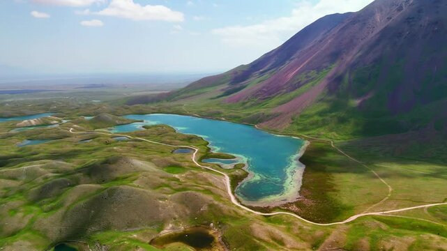 Aerial view of Tulpar Lake surrounded by green rolling hills and a purple mountain under a blue sky in Alay District, Osh Region, Kyrgyzstan.