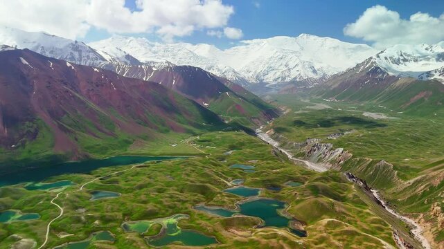 Aerial view of Tulpar Lake surrounded by green hills and red mountains with snow-capped peaks in the background in Alay District, Osh Region, Kyrgyzstan.