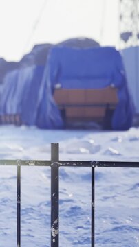 Snow covers the ground at a remote location. Structures are visible in the background, covered with blue tarps. The area looks quiet and empty under the clear sky.