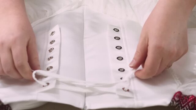 Clothes tying. Dressmaker work. Seamstress hands inserting string into lace loops of white embroidered corset. Fashion business workflow.