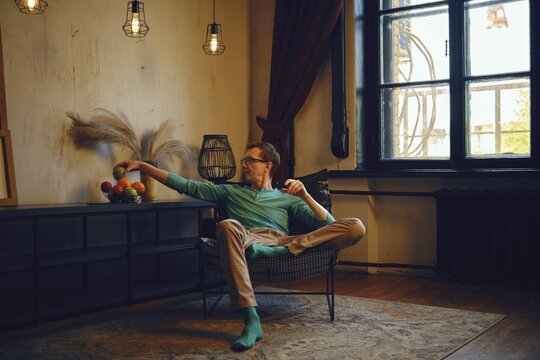 A man sits comfortably in a contemporary chair, casually extending his arm to grab fruit from a nearby table