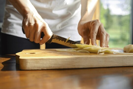 Unrecognizable person skillfully slices fresh ginger on a wooden cutting board in a well-lit kitchen