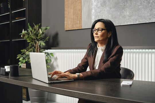 A businesswoman sits at a desk and typing on her laptop in an office. The space features elegant decor, including a plant and a minimalist color palette, enhancing productivity
