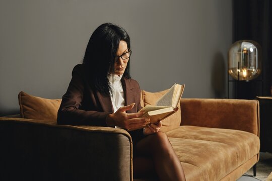 A person with glasses is deeply engrossed in reading a book while seated on a stylish couch in a contemporary living room. The ambient lighting casts a warm glow, creating a cozy atmosphere