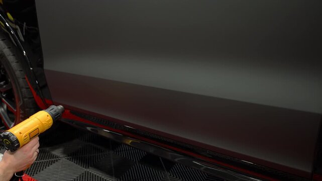Close up view of a heating matte vinyl wrap on a car body panel in a workshop. Automotive detailing process with film installation, smooth matte finish and precision work