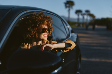 Fototapeta premium Woman with curly hair leans from car window, wearing sunglasses, enjoying a sunny drive, retro film color, palm trees in the background.