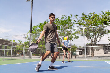 Fototapeta premium Pickleball doubles team playing active match on sunny outdoor court. Man moving with paddle while woman hitting yellow ball for healthy fitness.