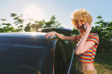 Fototapeta premium A woman in a striped crop top stands beside an open car window in bright sun, talking on a phone. Outdoor casual scene with vibrant colors conveys summer lifestyle, energy, and a cheerful road trip
