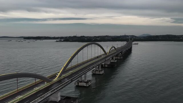 Aerial view of Dompak Bridge with its yellow arches spanning across the sea under a cloudy sky in Riau Islands, Indonesia.