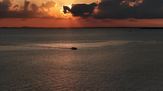 Aerial view of a small boat sailing across the calm sea during a golden sunset with dramatic clouds over Riau Island, Indonesia.