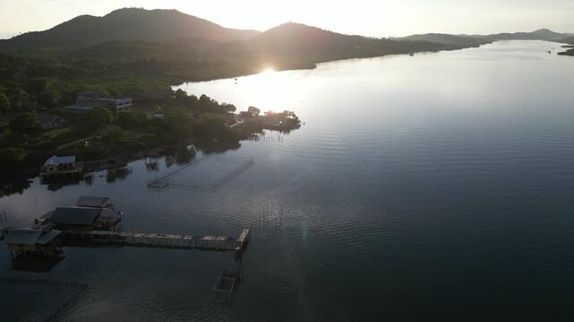 Aerial view of a coastal village with wooden stilt houses and piers along a calm river under the golden sun in Riau Islands, Indonesia.