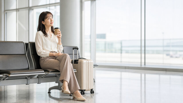 Japanese businesswoman sitting in airport waiting area with coffee and carry on suitcase