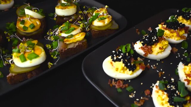Delicious canapes and deviled eggs topped with black caviar, bacon, and chives rotating on black plates, representing a luxurious and appetizing assortment of homemade party food