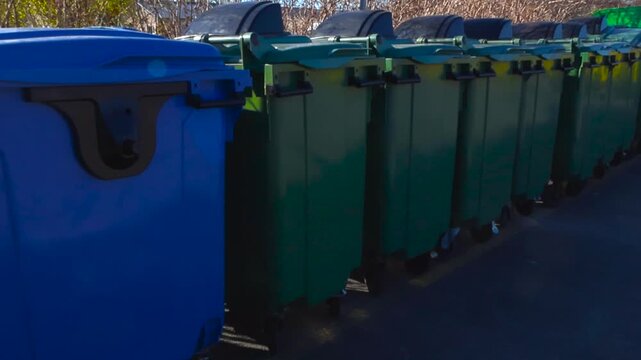 Numerous green and blue plastic garbage bins standing in a neat row outside on a sunny day, ready for waste collection and recycling, highlighting urban sanitation and environmental concepts