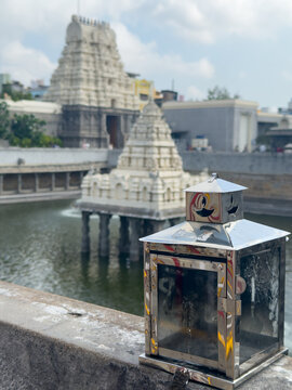 Metal diya stand at Kanchi Kamakshi Amman Temple tank in Kanchipuram, Tamil Nadu, with white shrine pavilion and gopuram backdrop.