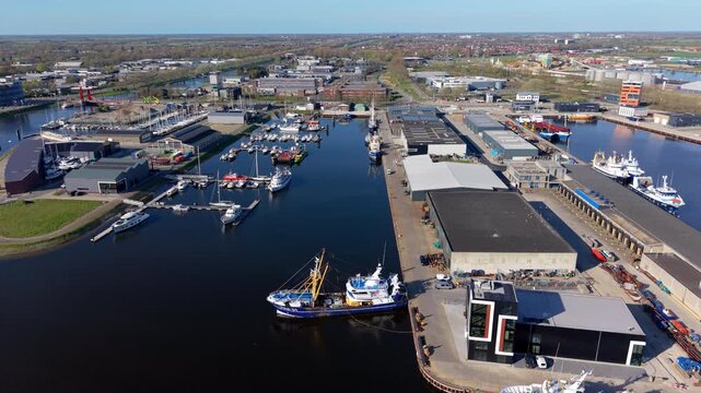 An aerial drone panorama of a bustling maritime port and industrial harbor, featuring docked fishing trawlers, yachts, marinas, warehouses, and an urban city landscape under a clear blue sky.