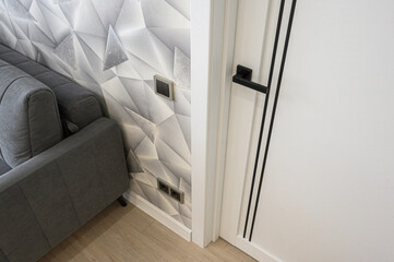 modern interior corner with a gray upholstered sofa, a white 3D geometric wall panel, and a minimalist white door with black vertical lines and handle © evgeniykleymenov