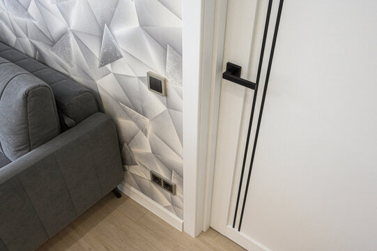 modern interior corner with a gray upholstered sofa, a white 3D geometric wall panel, and a minimalist white door with black vertical lines and handle