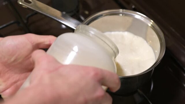 Man pours fresh milk into a metal saucepan on a gas stove. The process of heating milk in a steel pan in the kitchen, pouring milk from a glass jar into a saucepan and lighting a fire