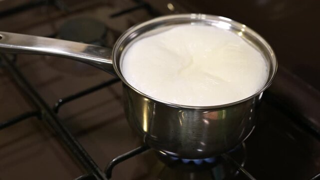Milk boiling over from a saucepan on a black gas stove burner. The moment of pouring hot foam over the edge of a steel pan, symbolizing the boiling of milk, not keeping track of the situation