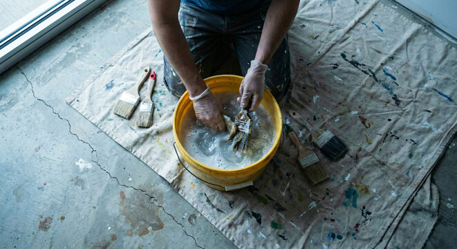 Overhead view of person kneeling on drop cloth cleaning paint brushes in a yellow bucket of soapy water