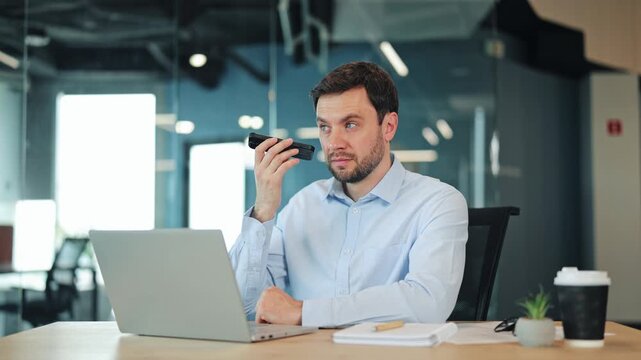 Focused businessman or IT specialist listening to audio note on cellphone. Corporate worker, manager or freelance designer using speakerphone or mobile app at contemporary coworking workspace.