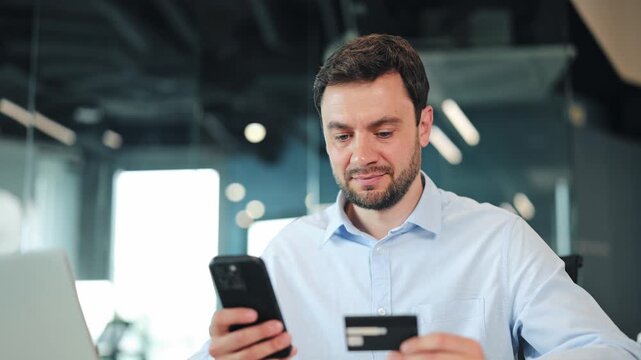 Happy businessman or IT specialist entering credit card number on smartphone and smiling at camera. Satisfied corporate worker, manager or freelance designer making online payment at office desk.