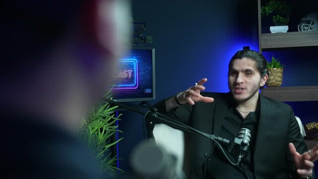 Young businessman slow motion gesturing while speaking into a microphone during a live interview in a podcast studio. Over the shoulder view of an audio broadcast recording and talk show concept
