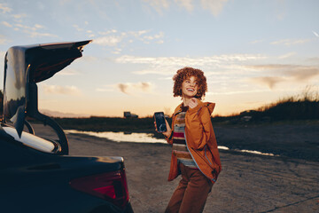 Fototapeta premium Woman lifestyle portrait outdoors at sunset, standing on a road by a parked car with an open trunk, curly haired woman holds a bag, travel mood, warm golden light