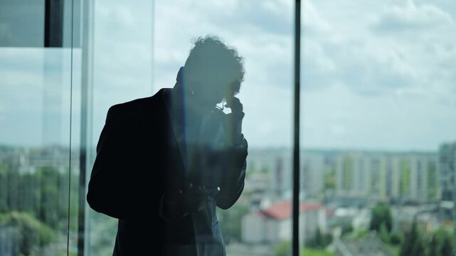 Professional silhouette of man standing at office window. Person holds smartphone while reading bad news, face shows deep worry and frustration about professional failure at the workplace right now.